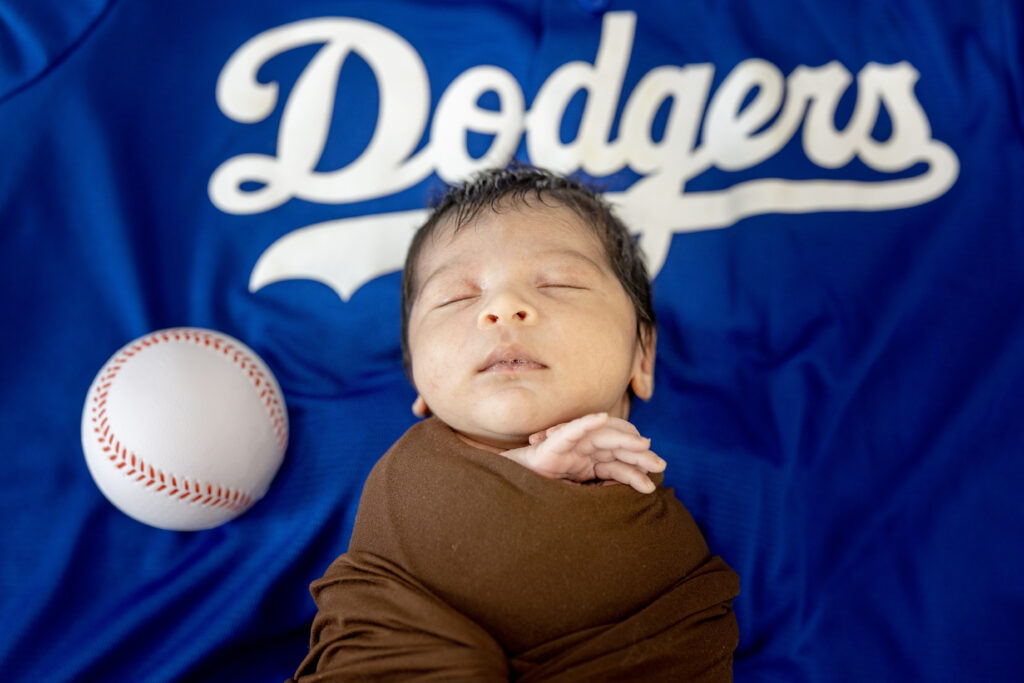 Baby boy lying on Dodgers-blue blanket with baseball props, professional newborn sports portrait.