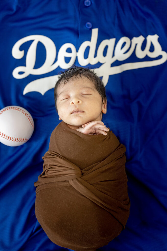 Swaddled newborn resting on blue Dodgers blanket, holding a tiny baseball.