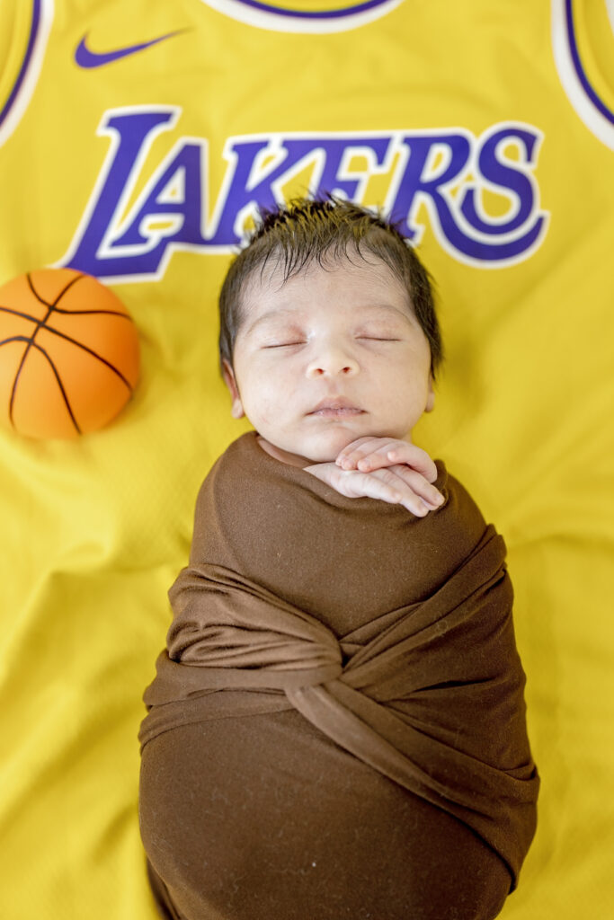 Infant wrapped in brown blanket surrounded by Lakers colors, Los Angeles team-inspired session.