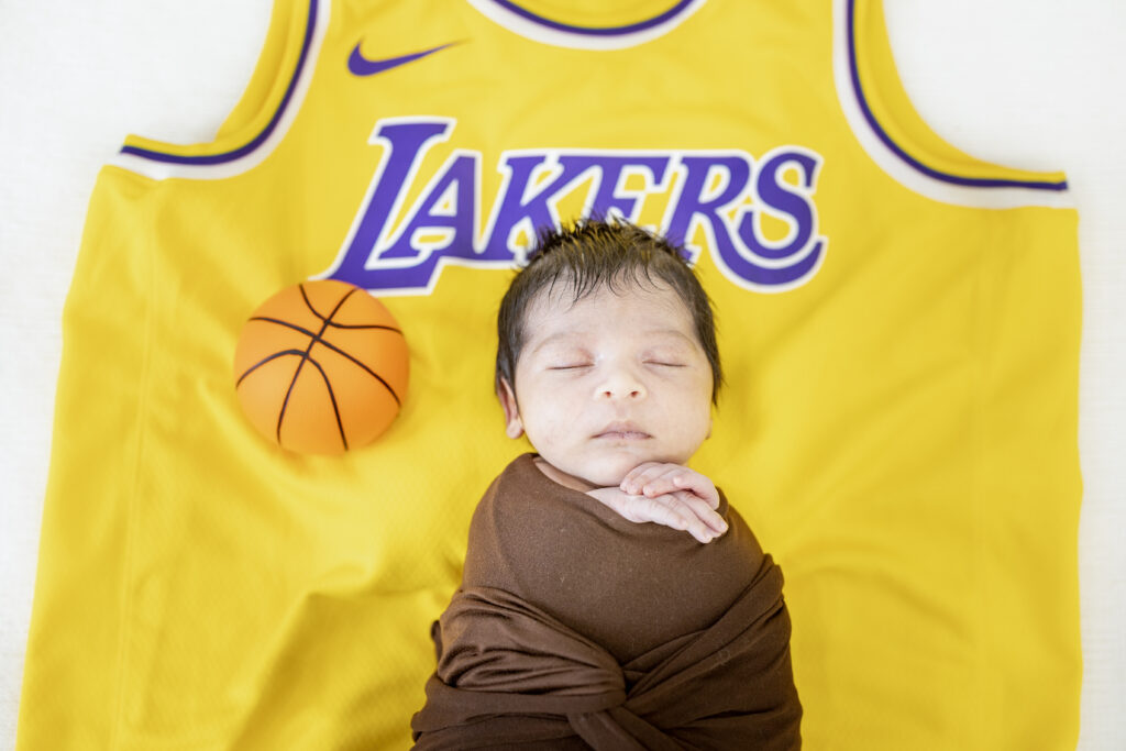 Newborn boy wrapped in soft yellow fabric, resting on Lakers jersey for themed photo shoot.