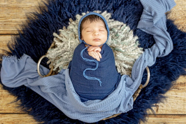 Full-length portrait of newborn boy wrapped in navy blue fabric, resting on fur rug.