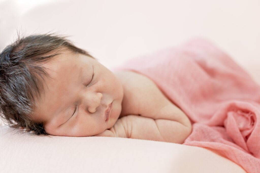 Newborn girl wrapped in soft pink blanket, sleeping peacefully on her side.