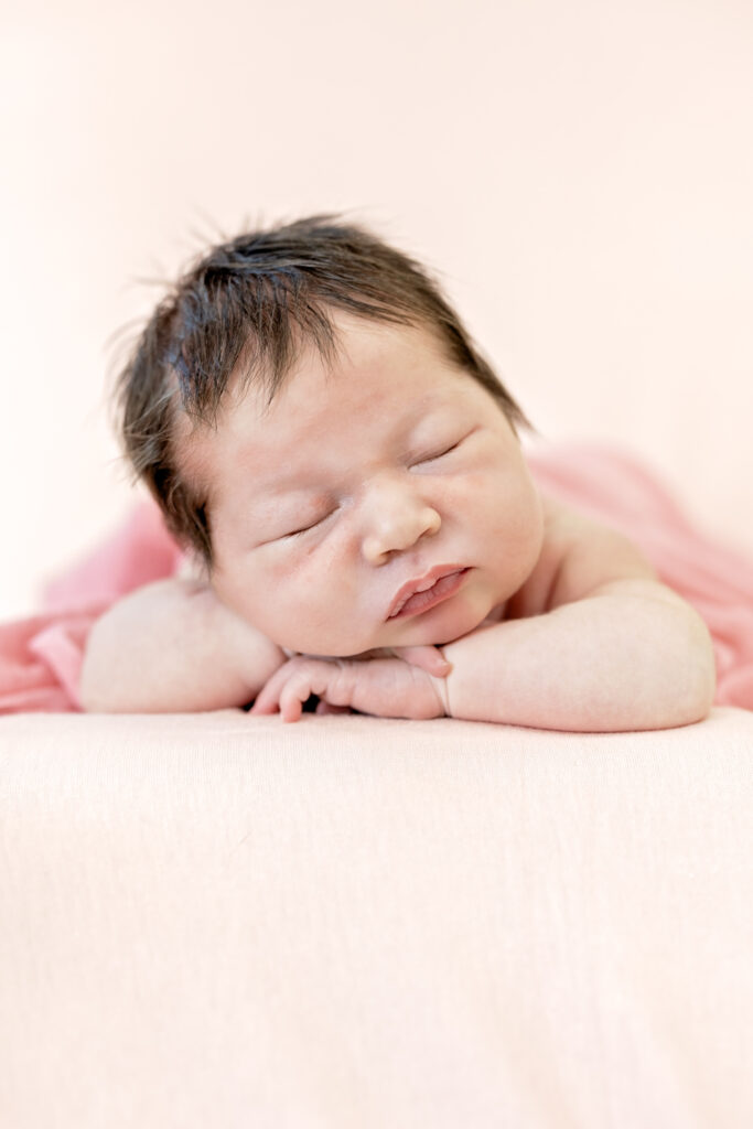 Newborn girl in pink wrap with floral headband, posed against soft pink background.