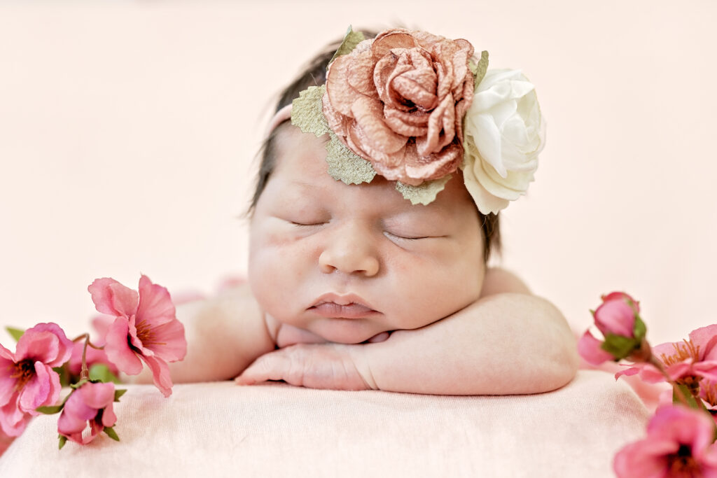 Newborn curled up on pink blanket surrounded by matching flowers and greenery.