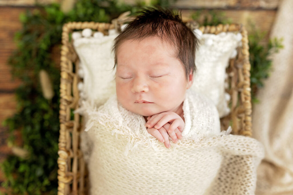 Cozy newborn in cream wrap inside a rustic basket on wooden floor