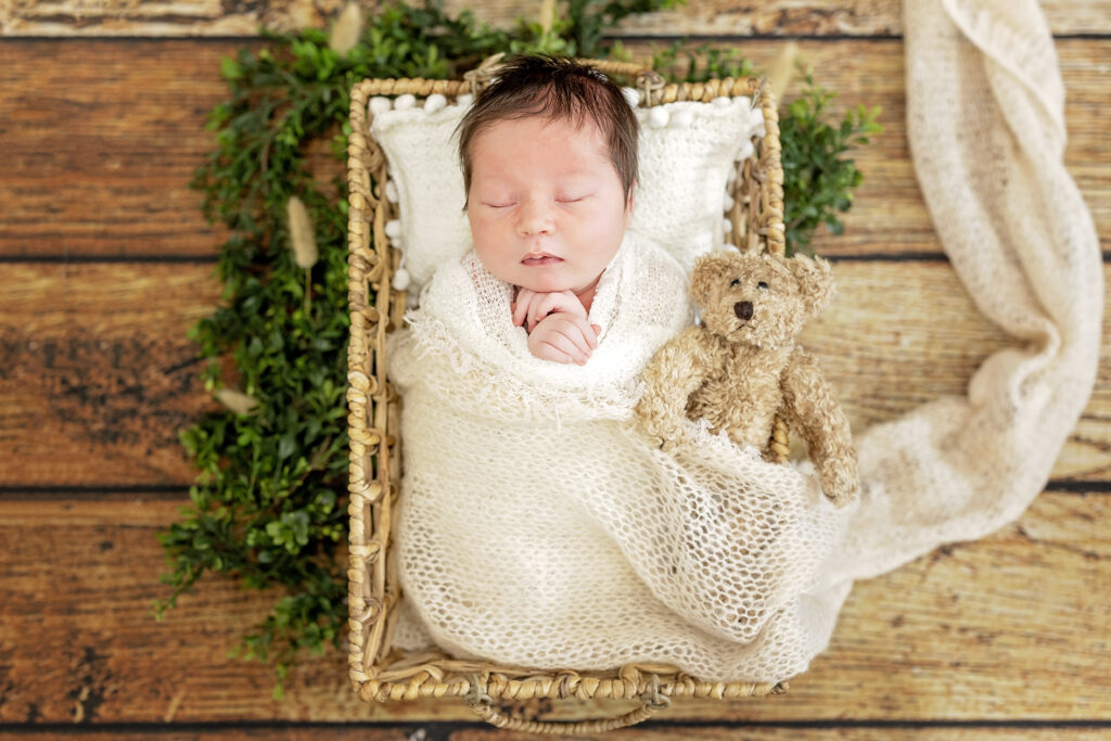 Overhead photo of baby in basket, ivory wrap, and greenery accent.