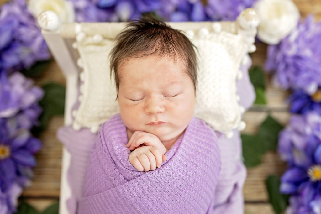 Baby girl surrounded by purple flowers and lavender wrap, bright floral newborn setup.