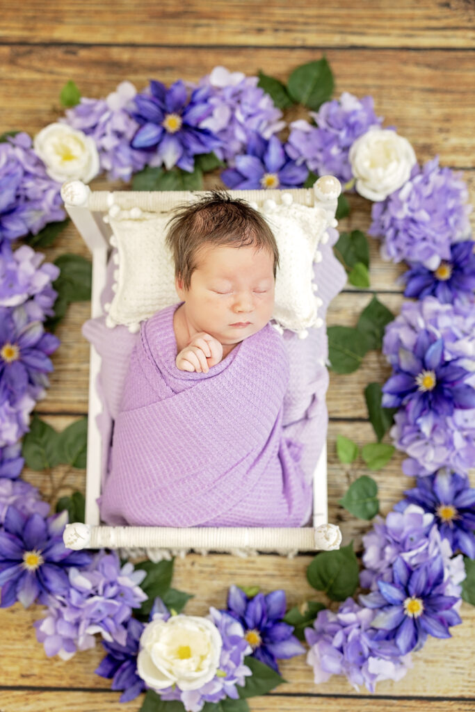 Wide floral arrangement framing sleeping baby girl in purple tones.