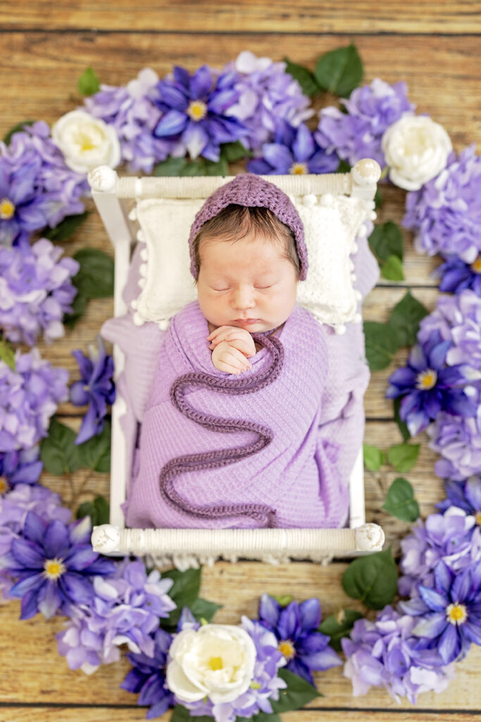 Baby wrapped in lavender fabric surrounded by purple florals, rustic floor background.