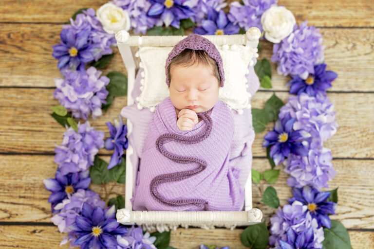 Newborn swaddled in deep purple wrap, nestled within floral ring.