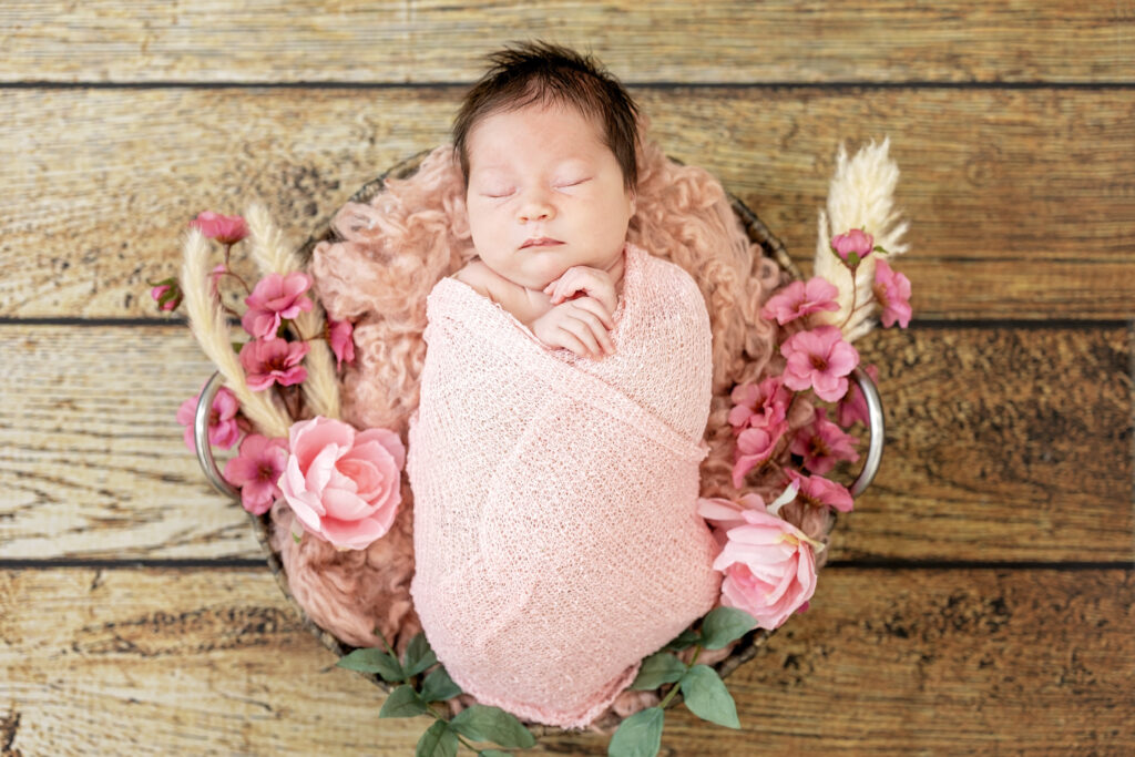 Smiling newborn girl in pink wrap, posed on wooden floor with soft textures.