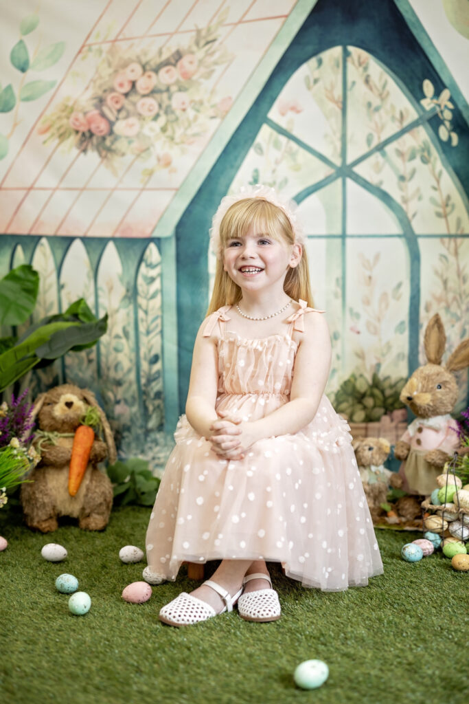 Child smiling during an Easter mini photo session, seated in a pastel spring greenhouse backdrop with Easter bunnies, flowers, and decorative eggs.