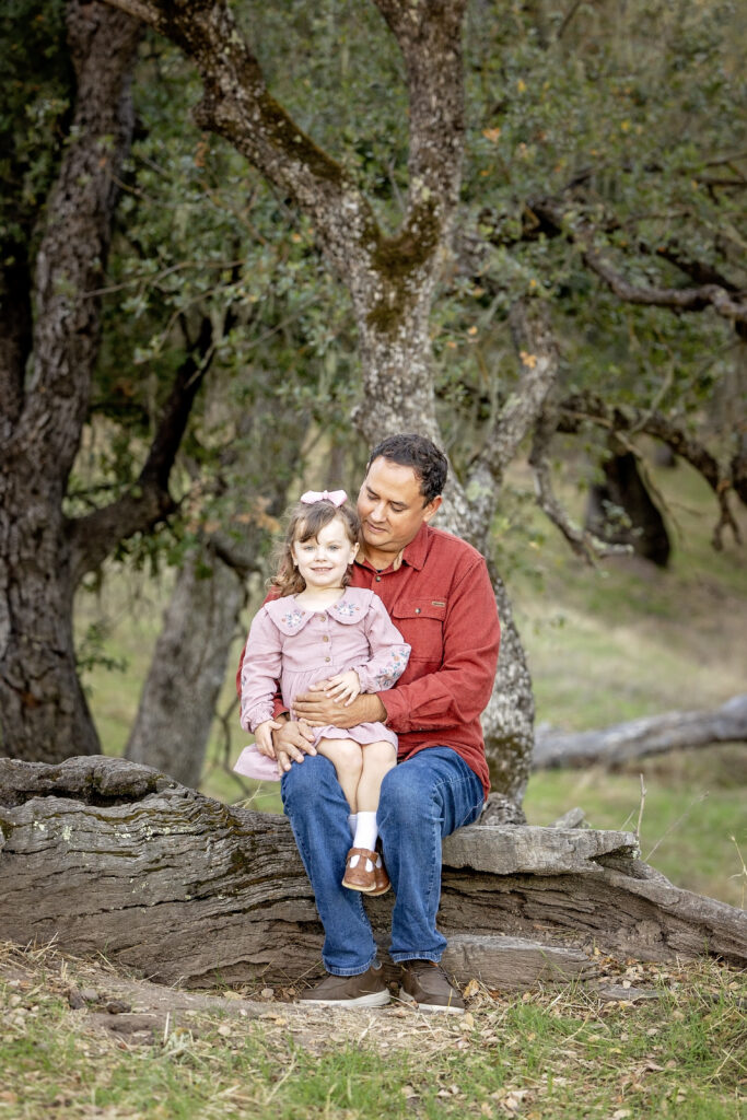 Dad sitting on a fallen tree with daughter on his lap, both smiling softly in a wooded area