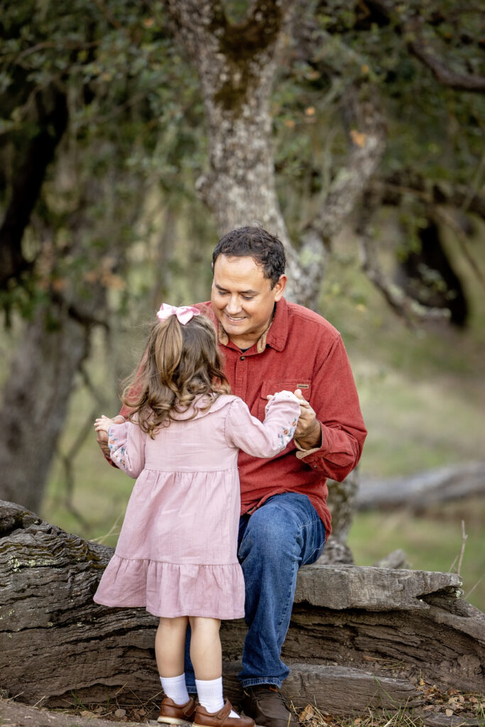 Dad smiling at daughter while holding her hands as she faces him during their outdoor session
