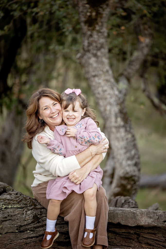 Mom hugging daughter tightly while sitting on a log, both smiling at the camera with trees behind them