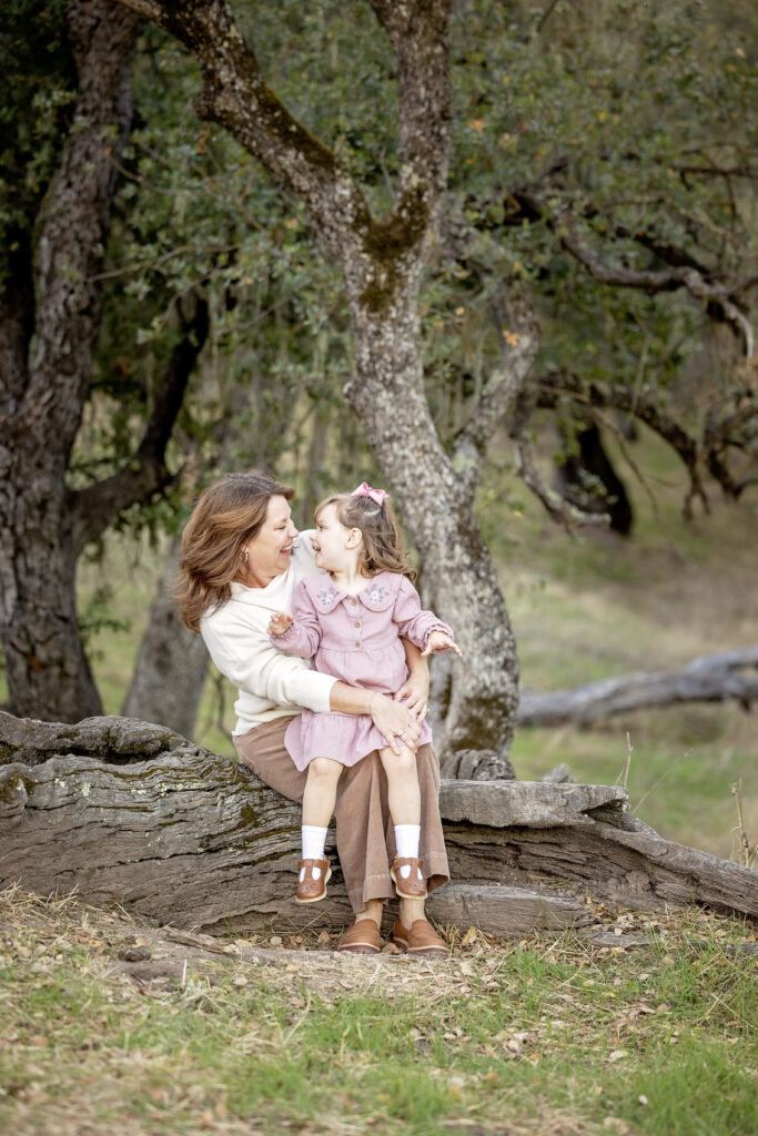 Mom and daughter sitting together on a fallen tree in the woods, looking at each other and smiling