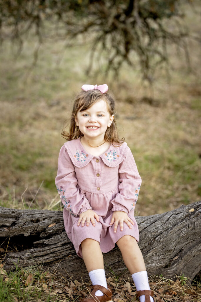Girl sitting on a log, hands in lap, smiling widely during her family photo session