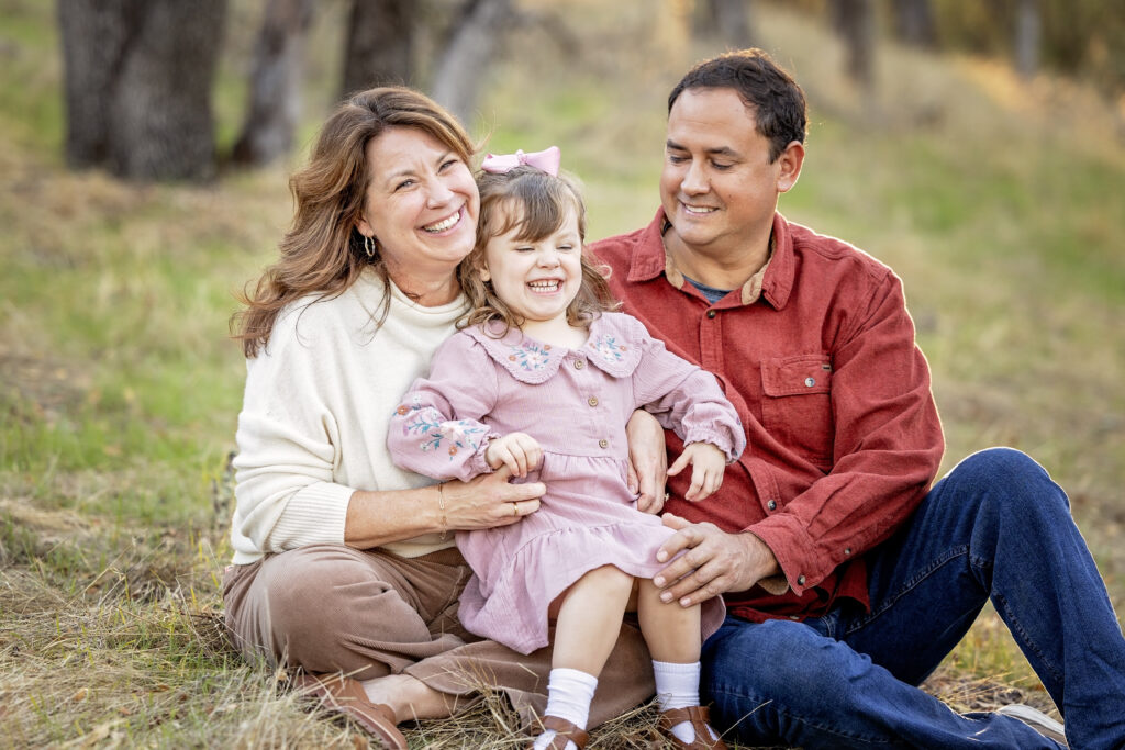Family of three sitting together in the grass, smiling and cuddling during their golden hour session in Atascadero