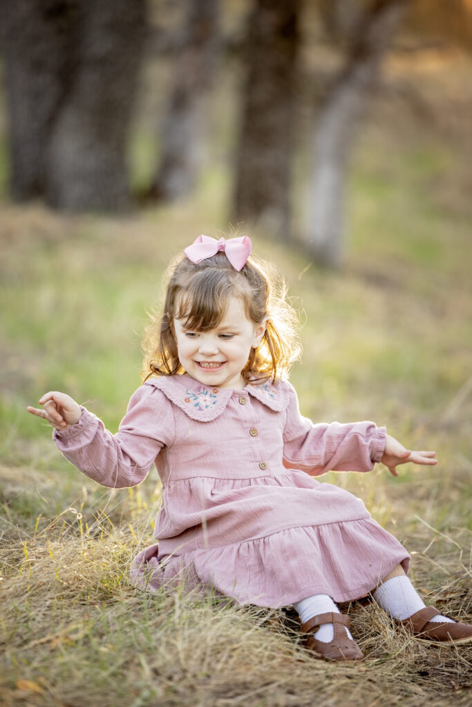 Young girl in a pink dress sitting in the grass and laughing during a sunset photo session at Jim Green Trail in Atascadero