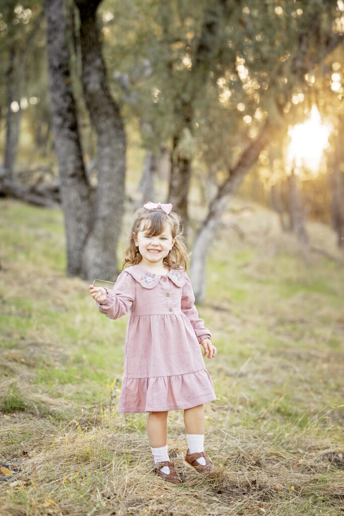 Little girl in a pink dress walking through the trees at sunset with a playful expression