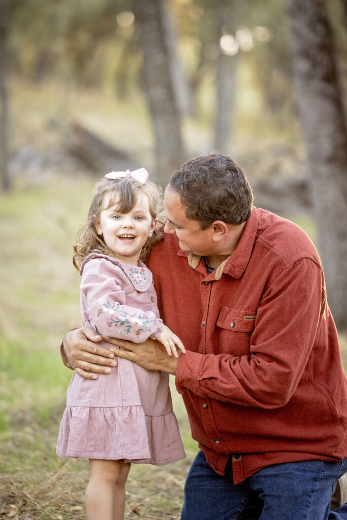 Dad playfully kissing his daughter's cheek as she giggles, surrounded by trees